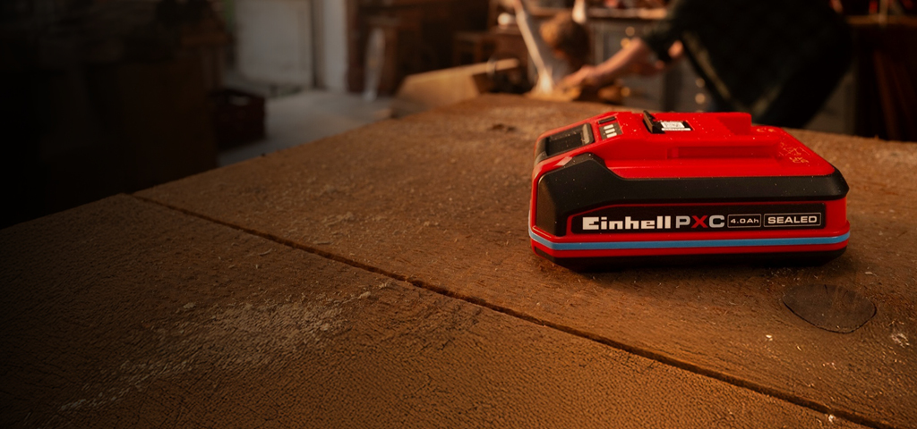 Batería Einhell Power X-Change 4.0Ah SELLADA en un banco de trabajo de madera, que muestra un anillo y una etiqueta azules. 