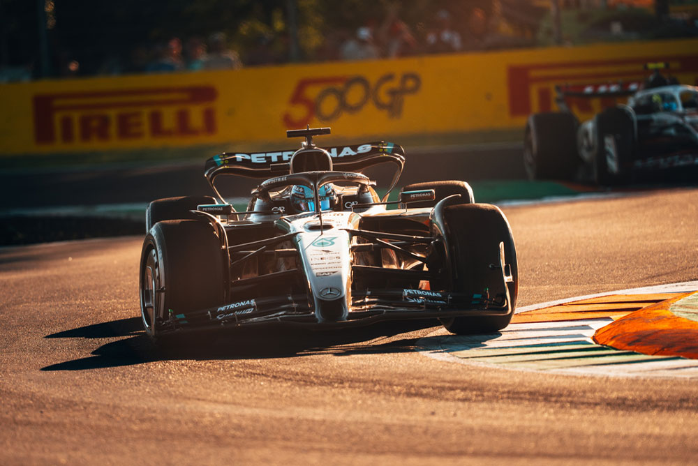A Mercedes-AMG PETRONAS Formula One car racing on a sunlit track during a Grand Prix. The car is taking a corner at high speed with another car in the background.
